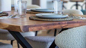Una mesa de comedor dispuesta con platos, vasos y cubiertos. Hay una planta sobre la mesa y se ve una cocina al fondo.