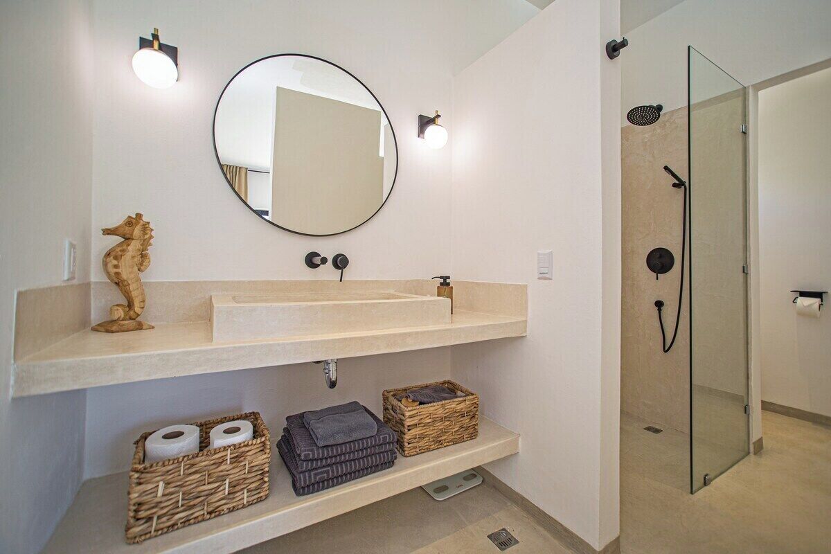 Un baño con un espejo redondo, un lavabo beige, una decoración con caballito de mar y una ducha con cerramiento de vidrio.