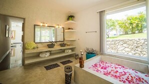 Casa de banho com uma janela grande, banheira com pétalas de rosa, espelho, prateleiras com produtos de higiene e uma planta em vaso.