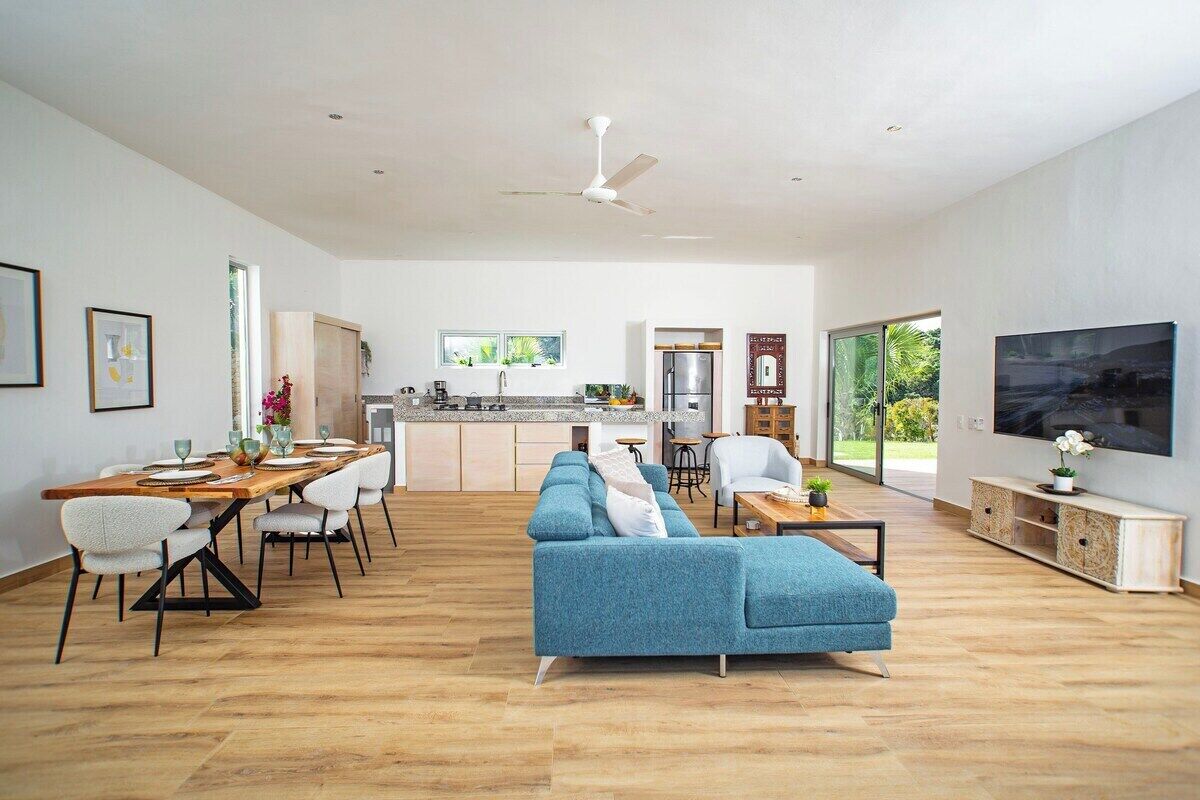 A spacious living area with a blue sofa, dining table, and chairs. A kitchen is visible in the background with a refrigerator and a window.