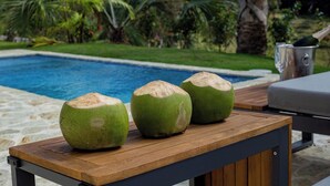Una mesa de madera con tres cocos encima, con vista a una piscina y un jardín tropical al fondo.