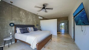 A bedroom with a bed, a ceiling fan, a TV, and a view of a beach through the window.