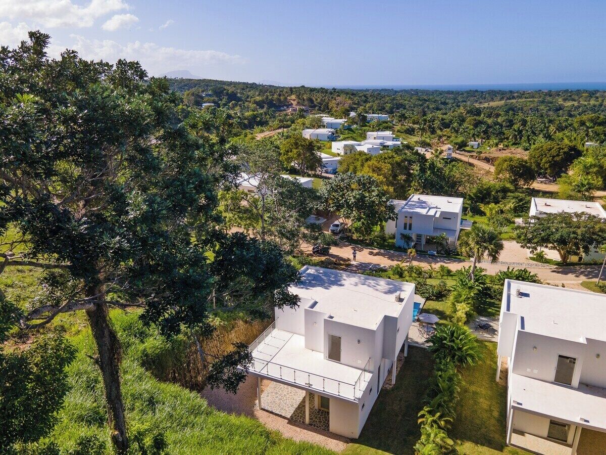 A residential area with modern white buildings, surrounded by lush greenery and a clear view of the ocean in the distance.