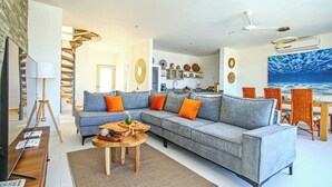Een moderne woonkamer met een grijze bank, een houten salontafel en een eetgedeelte met rieten stoelen.