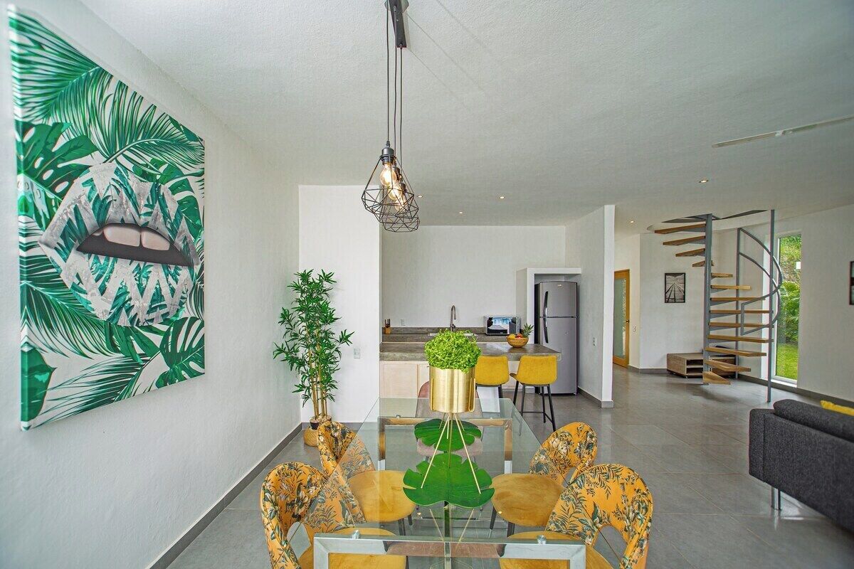 Een moderne woonkamer met een glazen eettafel, gele stoelen, een groot abstract schilderij, een trap en een keuken op de achtergrond.