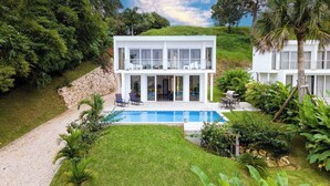 Une maison moderne à deux étages, dotée d’une piscine, entourée d’une végétation luxuriante et de palmiers.