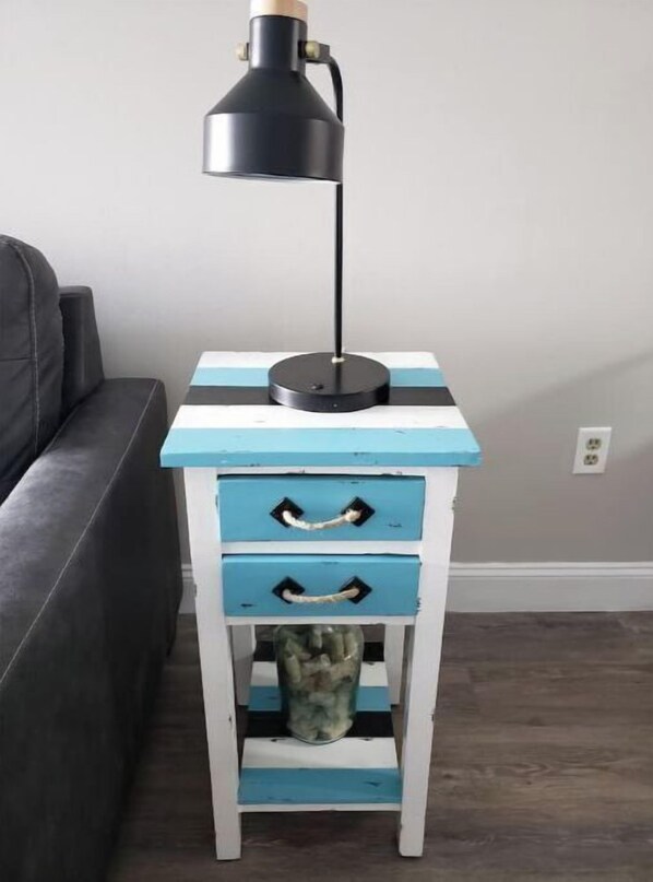 Een klein nachtkastje met twee lades, waarop een zwarte lamp staat, geplaatst tegen een muur in een kamer met een grijze bank.