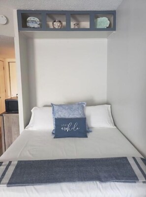 A bedroom with a bed, a pillow with text, a shelf above the bed, and a door to the left. - Cute Condo with Pool (Huddleston)