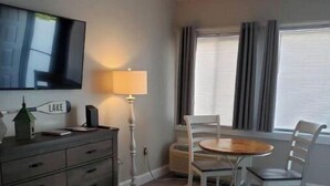 A hotel room with a flat-screen TV mounted on the wall, a wooden dresser with drawers, a small table with chairs, a lamp, and a window with curtains. - Cute Condo with Pool (Huddleston)