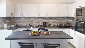 Une cuisine moderne avec des armoires blanches, un pare-feu gris et des appareils en acier inoxydable. On y trouve un coin repas avec deux tabourets de bar et une table dressée avec un panier de fruits et une assiette de nourriture.