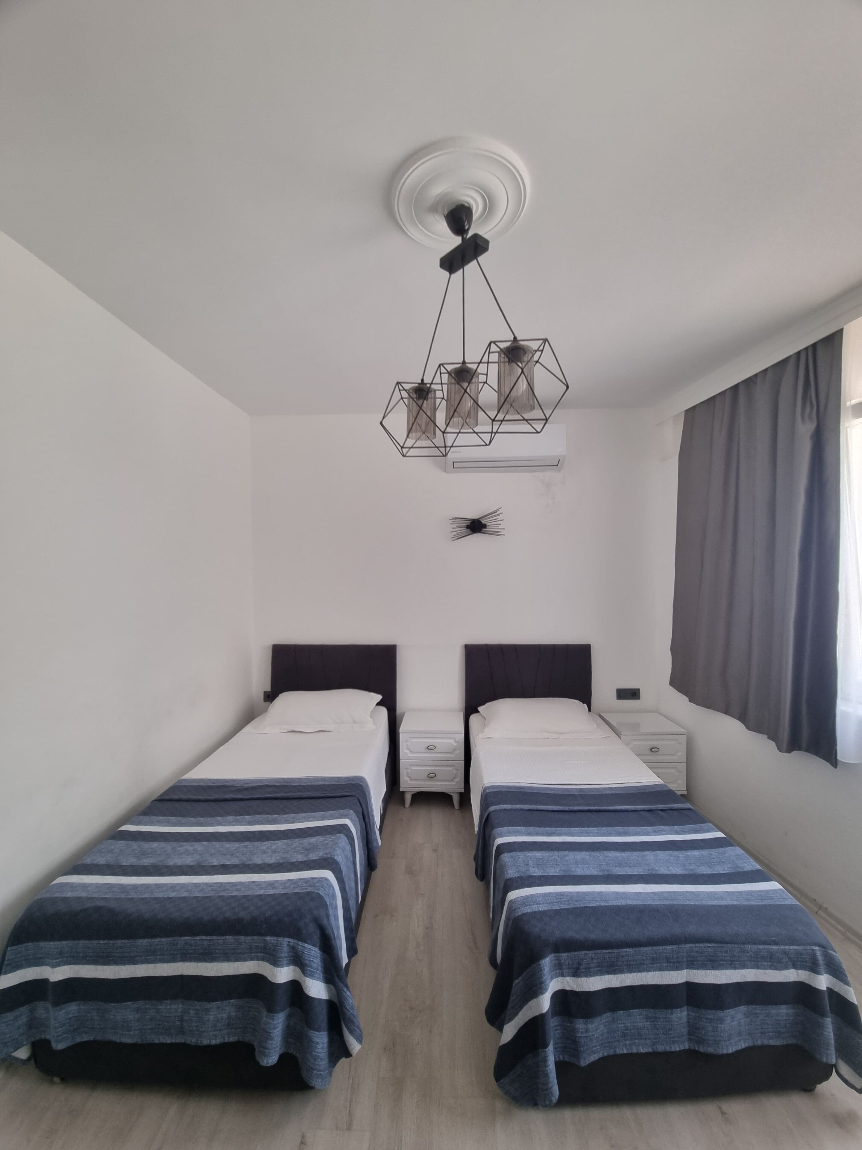 A room with two single beds, a small bedside table with a lamp, a ceiling light fixture, and a window with curtains.