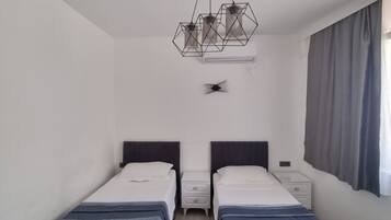 A room with two single beds, a small bedside table with a lamp, a ceiling light fixture, and a window with curtains.