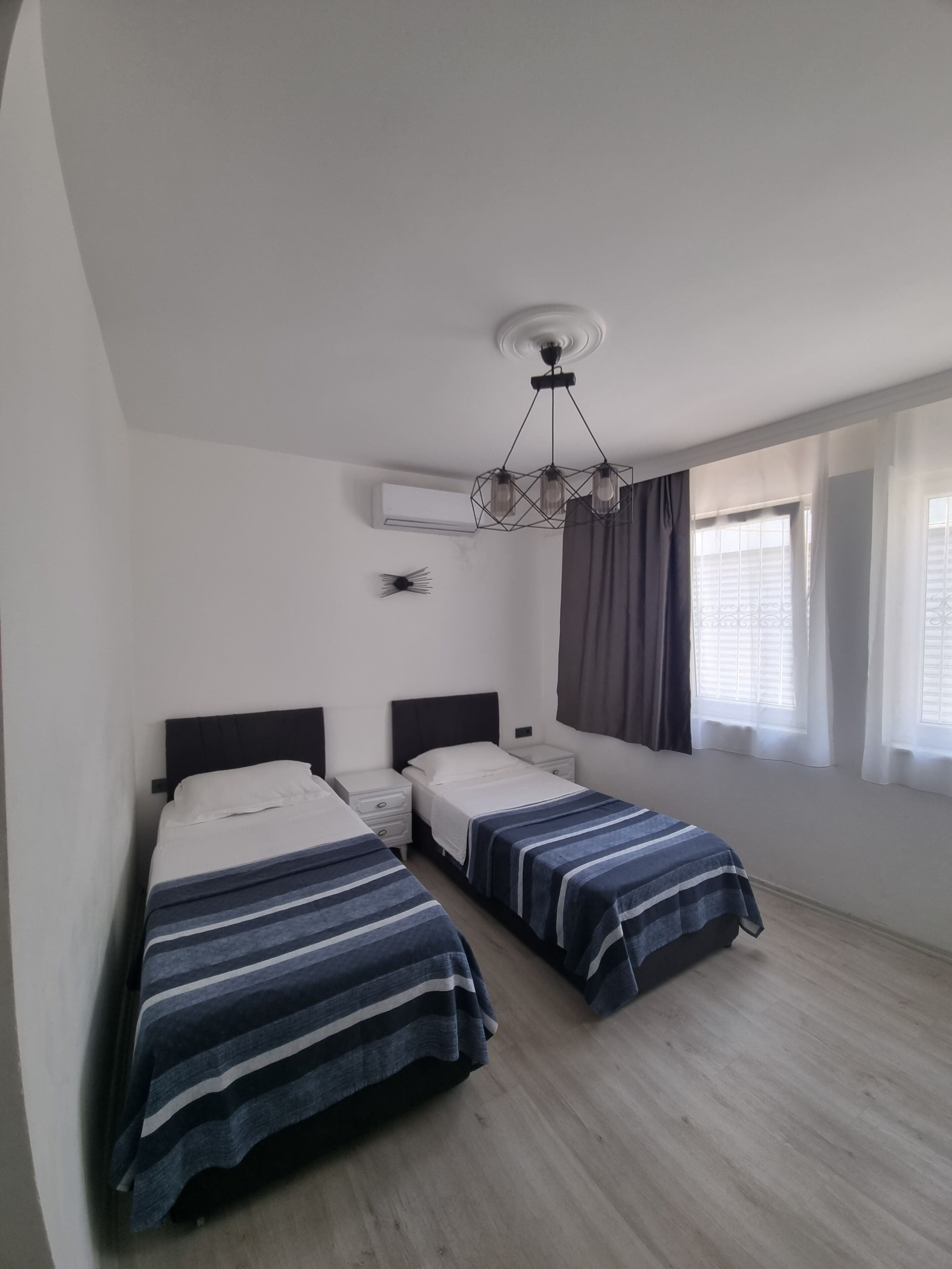 A bedroom with two beds, a modern chandelier, an air conditioning unit, and a window with curtains.