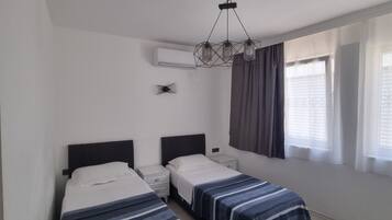 A bedroom with two beds, a modern chandelier, an air conditioning unit, and a window with curtains.