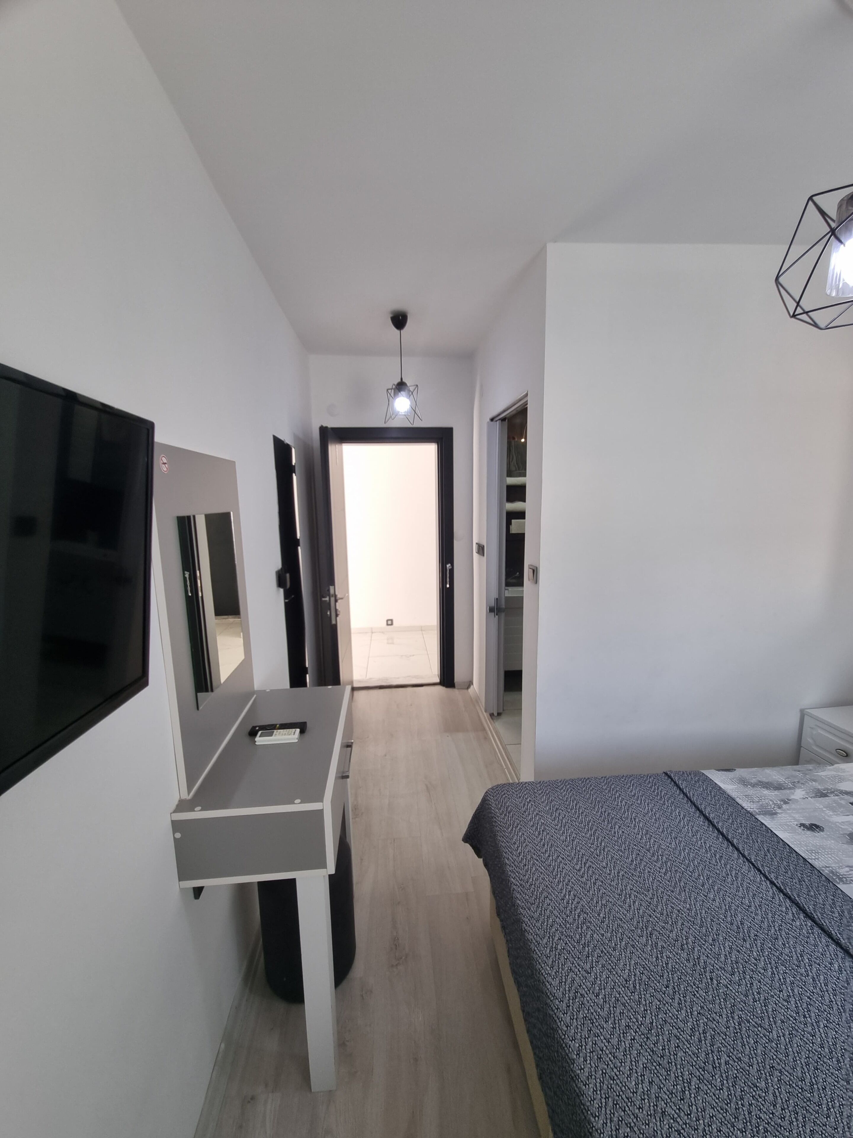 A modern bedroom with a bed, a desk with a chair, a TV mounted on the wall, and a door leading to a bathroom.