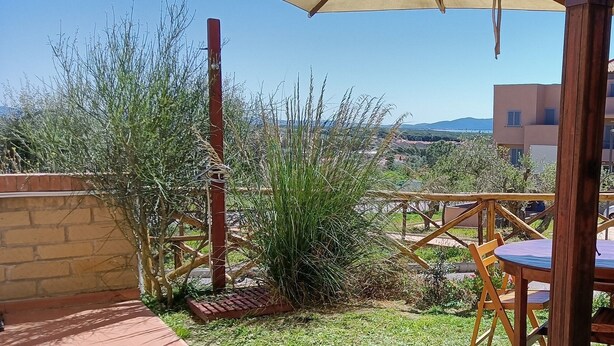 A patio area with a wooden table and chairs, a large umbrella, and a view of a distant town and hills. - Sea view apartment (Castiglione della Pescaia)