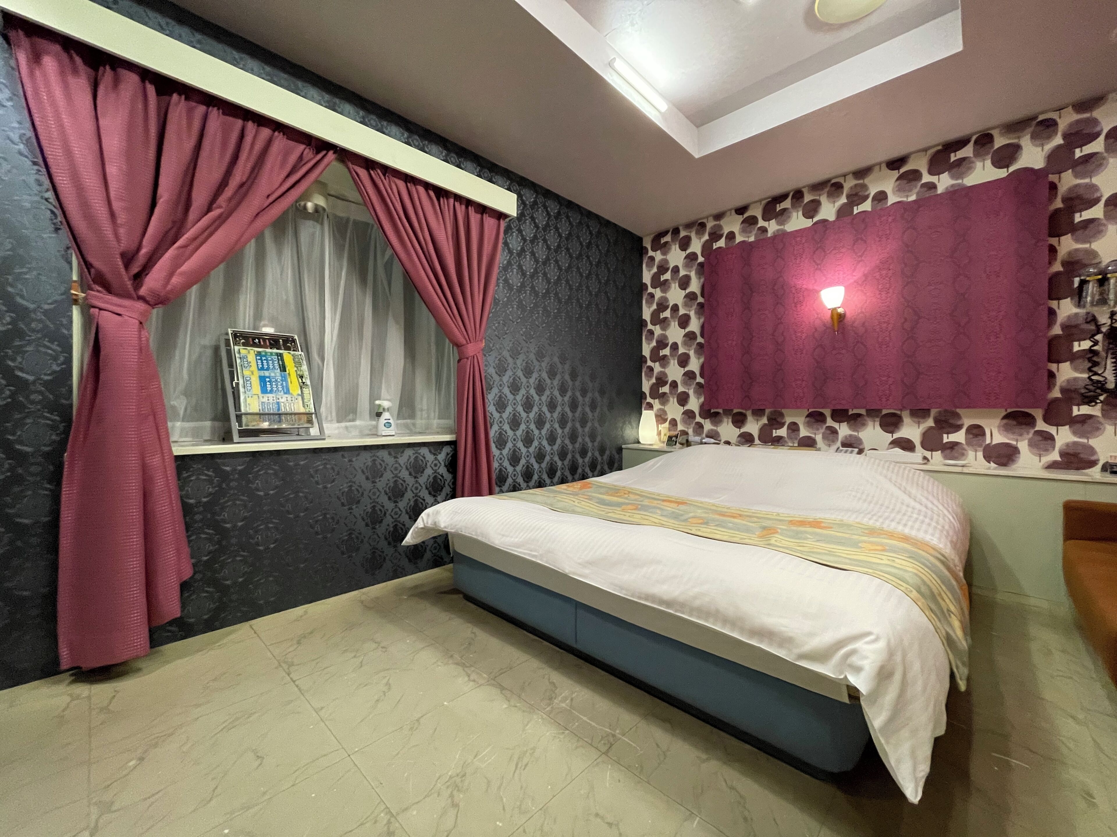 Quarto com cama, cortinas, janela, prateleira na parede com produtos de higiene pessoal e parede estampada.