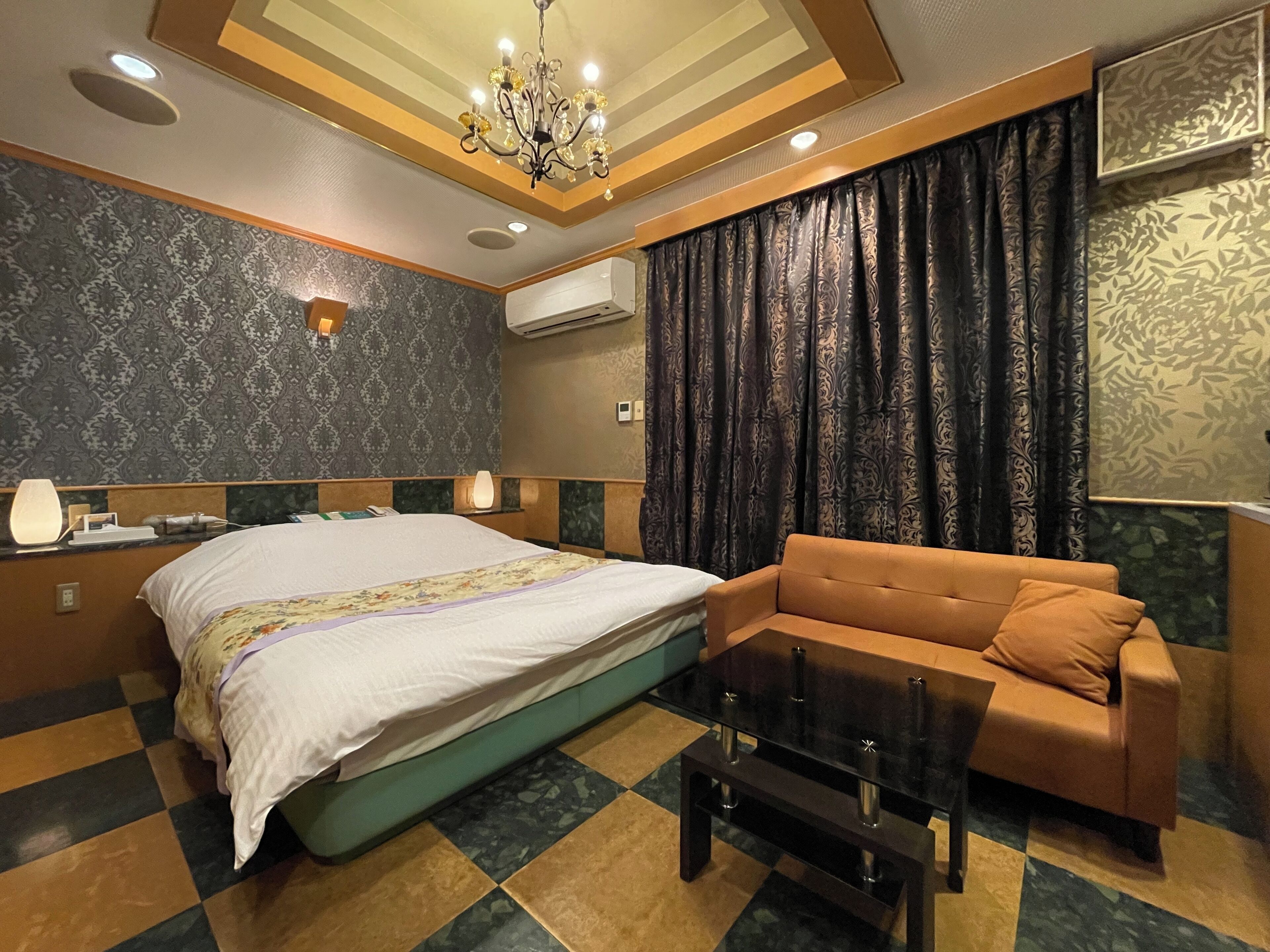 Een hotelkamer met een bed, een bank, een salontafel, een kroonluchter, behang met een patroon en een airconditioner.