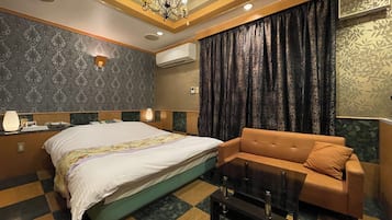 A hotel room with a bed, a sofa, a coffee table, a chandelier, patterned wallpaper, and an air conditioning unit.