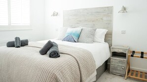 A neatly made bed with rolled towels, a wooden headboard, bedside tables, and a small wooden bench.