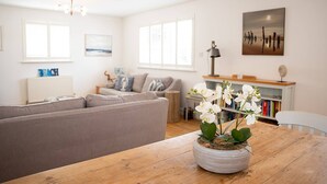 A modern living room with a grey sofa, a wooden dining table with a potted orchid, and a bookshelf in the background.