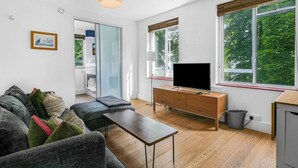 Una sala de estar moderna con un sofá, una mesa de centro, un televisor sobre un mueble de madera y ventanas amplias con vistas a zonas verdes.