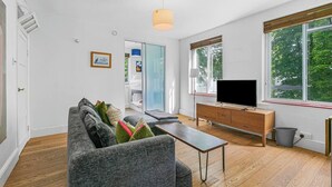 Una sala de estar moderna con un sofá gris, una mesa de centro de madera y un televisor sobre un mueble de madera. Se aprecia un ventanal con persianas, una puerta que da a otra habitación y una lámpara de techo.
