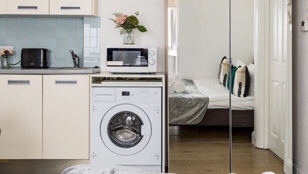 Une chambre d’hôtel compacte comprenant un coin cuisine, un lave-linge, un lit avec une couette grise et un miroir qui reflète la pièce.