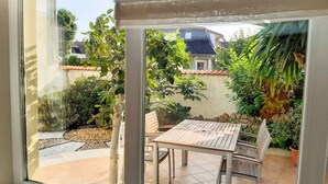 Uitzicht door een raam op een terras met een tafel en stoelen, omgeven door verschillende planten en een stenen muur.