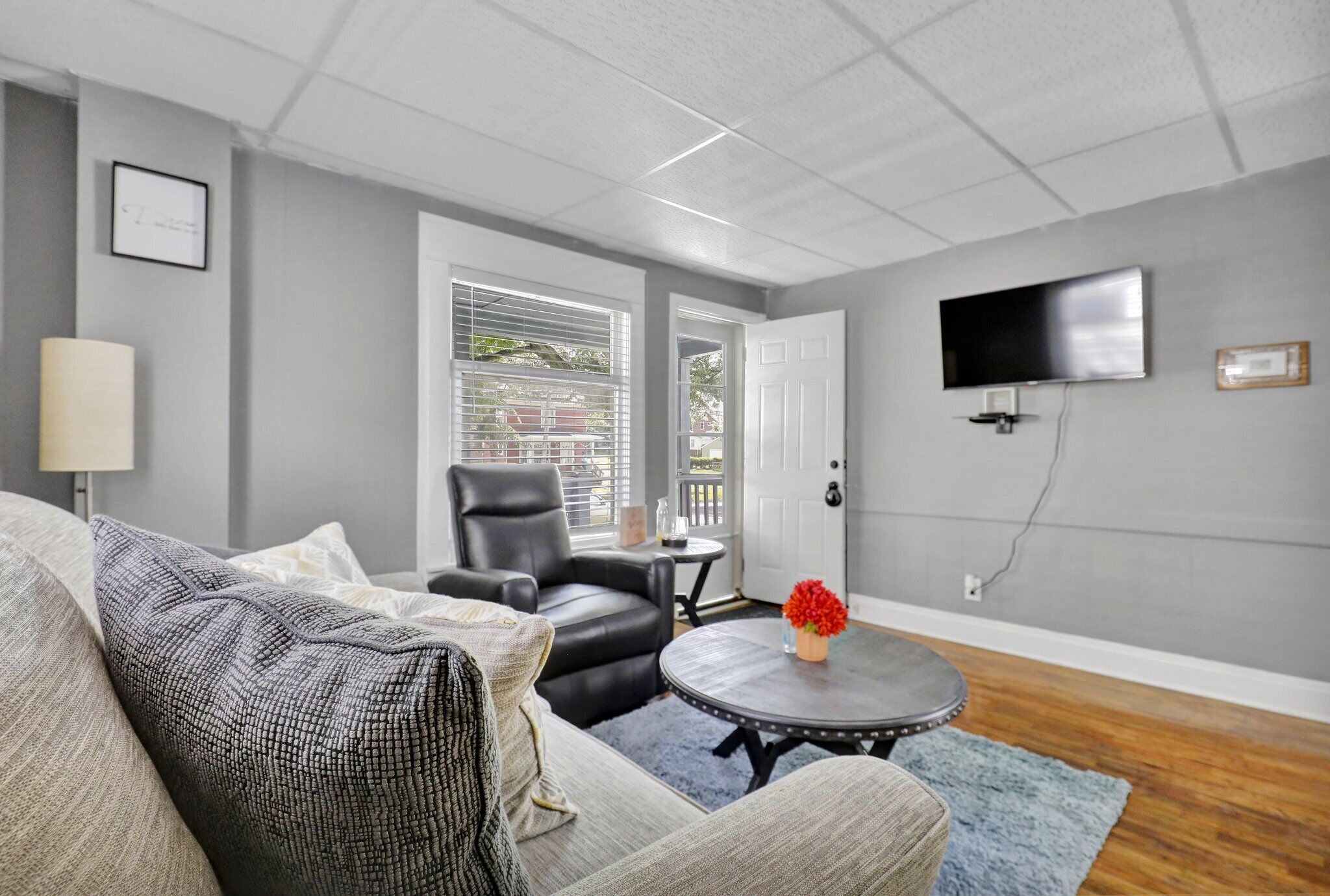 A living room with a grey couch, a black leather chair, a round coffee table with a red flower, and a flat-screen TV mounted on the wall.