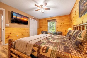 A bedroom with a wooden floor, a bed with a quilt, a ceiling fan, a TV on the wall, and a window with curtains. - π¬ Carefree Hideaway | Theater Room + Pet-Friendly + Hot Tub π (Sevierville)