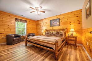 A log cabin bedroom with a wooden bed, two chairs, a ceiling fan, and a window with curtains.