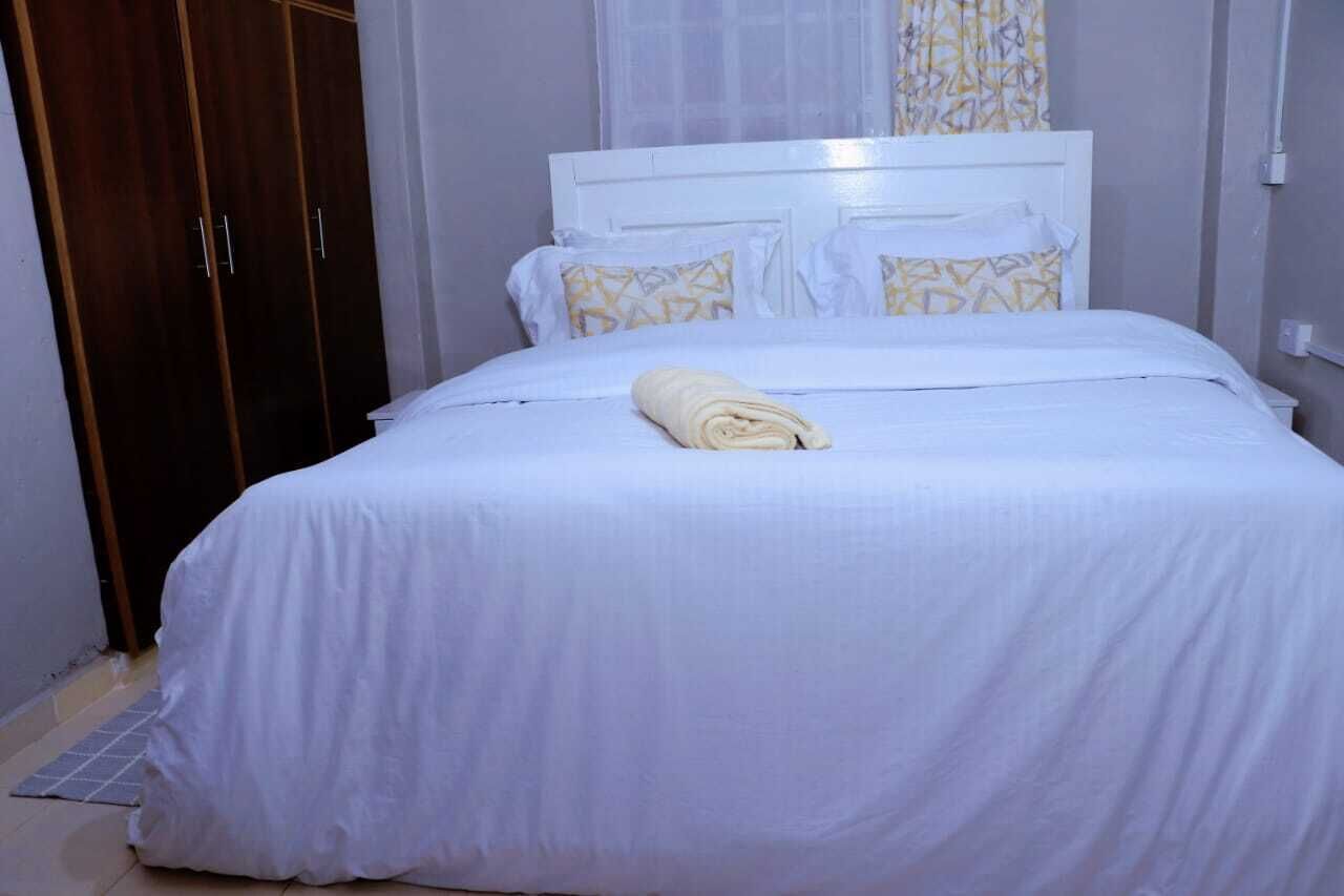 A neatly made bed with a white bedspread and pillows, a rolled towel on the bed, and a wooden wardrobe in the background.