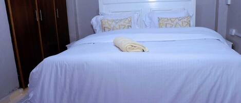 A neatly made bed with a white bedspread and pillows, a rolled towel on the bed, and a wooden wardrobe in the background.