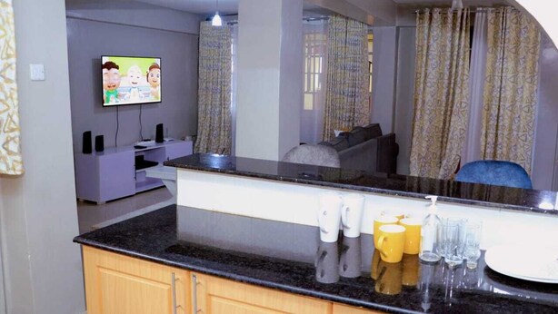 A modern kitchen with a granite countertop, wooden cabinets, a built-in oven, and a flat-screen TV mounted on the wall. - Lux Suites Okiega Apartments Kisii (Kisii)