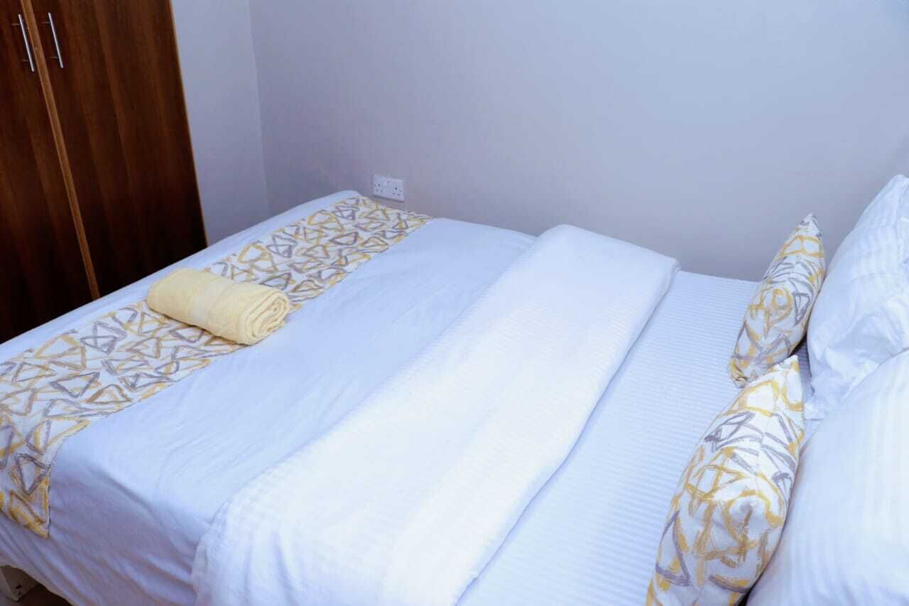 Een net opgemaakt bed met een bedspreidek en kussens, een houten garderobe en een witte muur met een stopcontact.