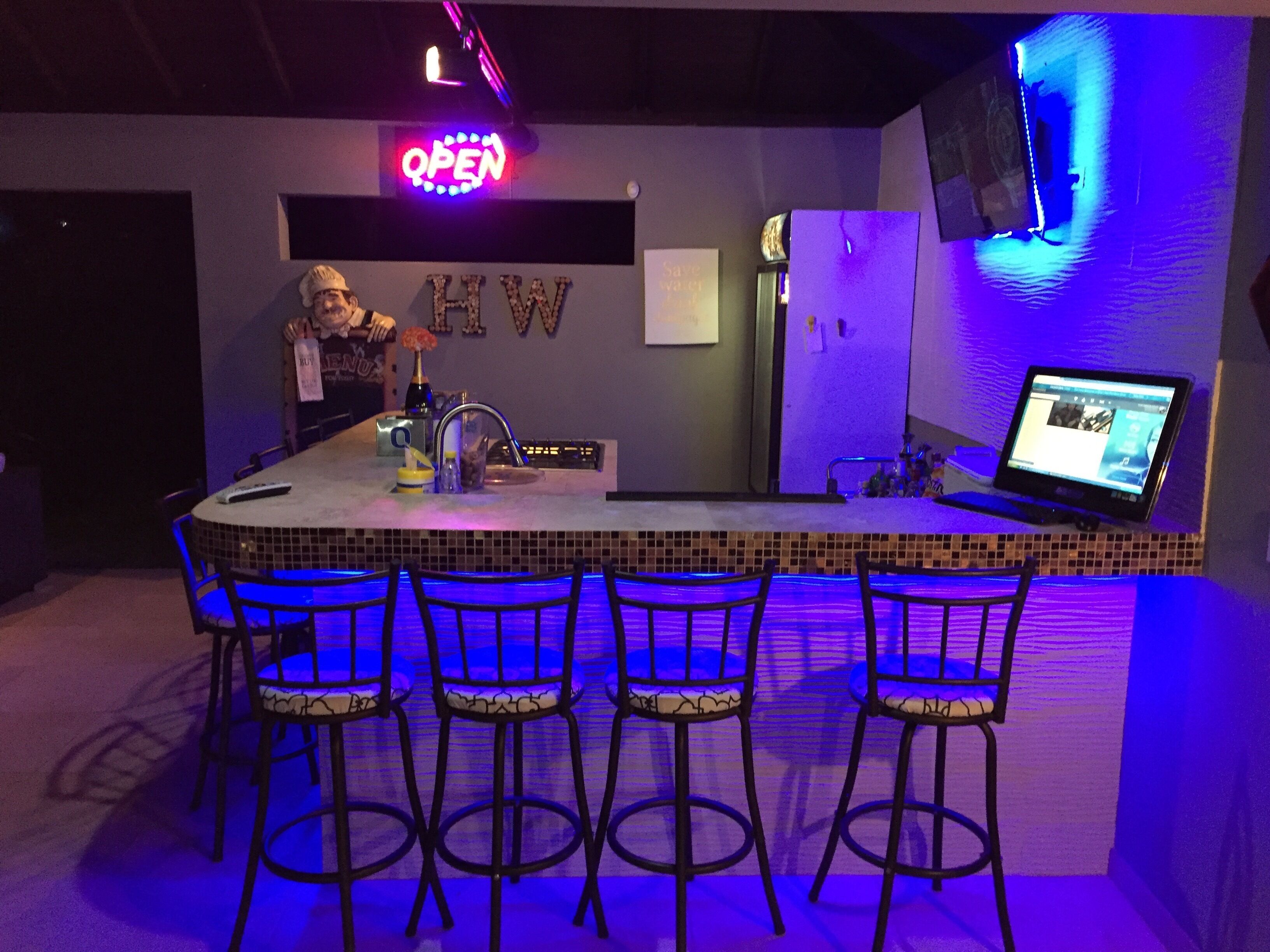 A bar area with a neon 'OPEN' sign, a 'H2O' sign, and a laptop on the counter.