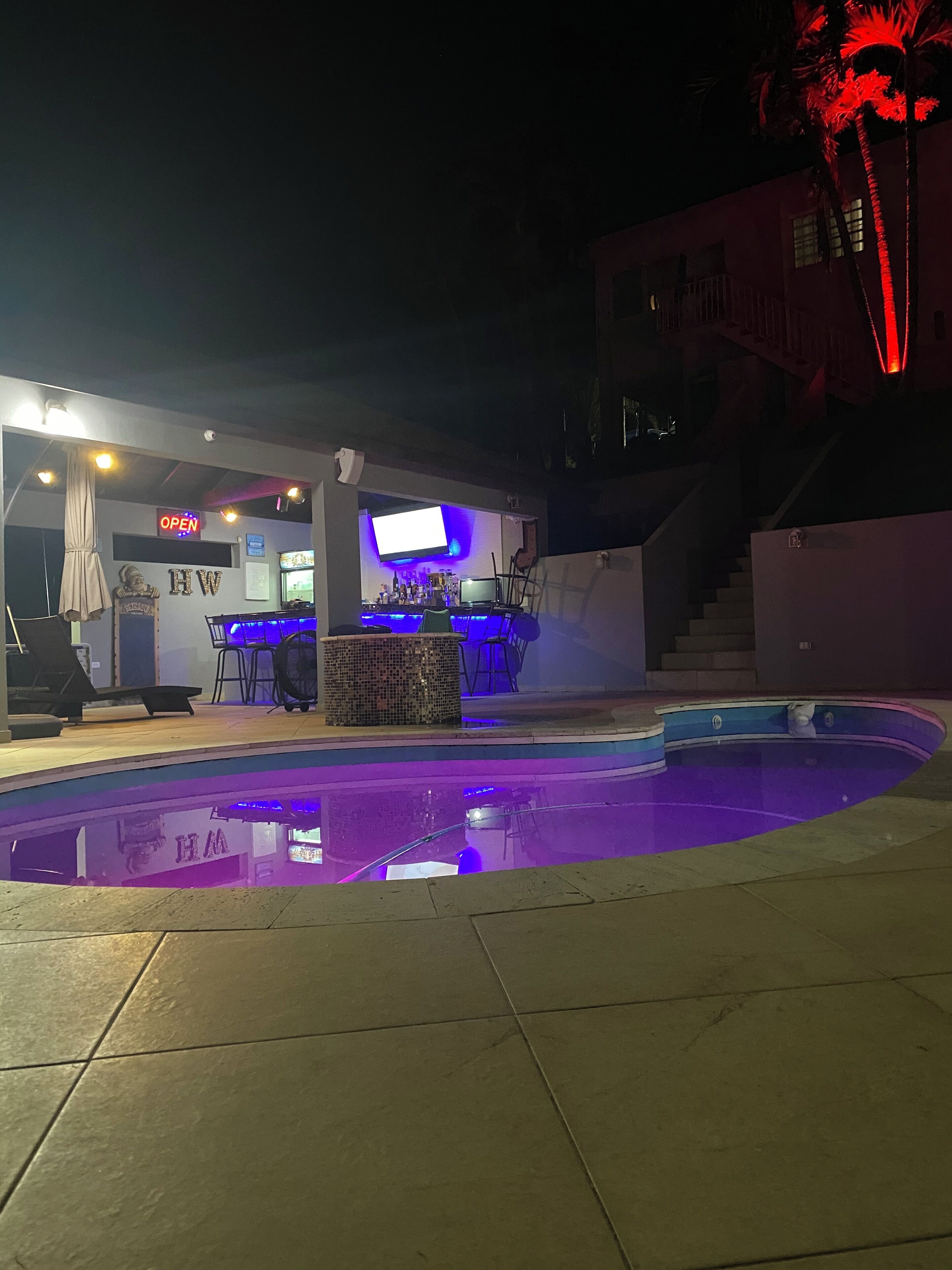 Ein Poolbereich mit Bar und Sitzgelegenheiten, ein Schild mit der Aufschrift 'OPEN' und eine Treppe, die zu einer erhöhten Ebene führt.