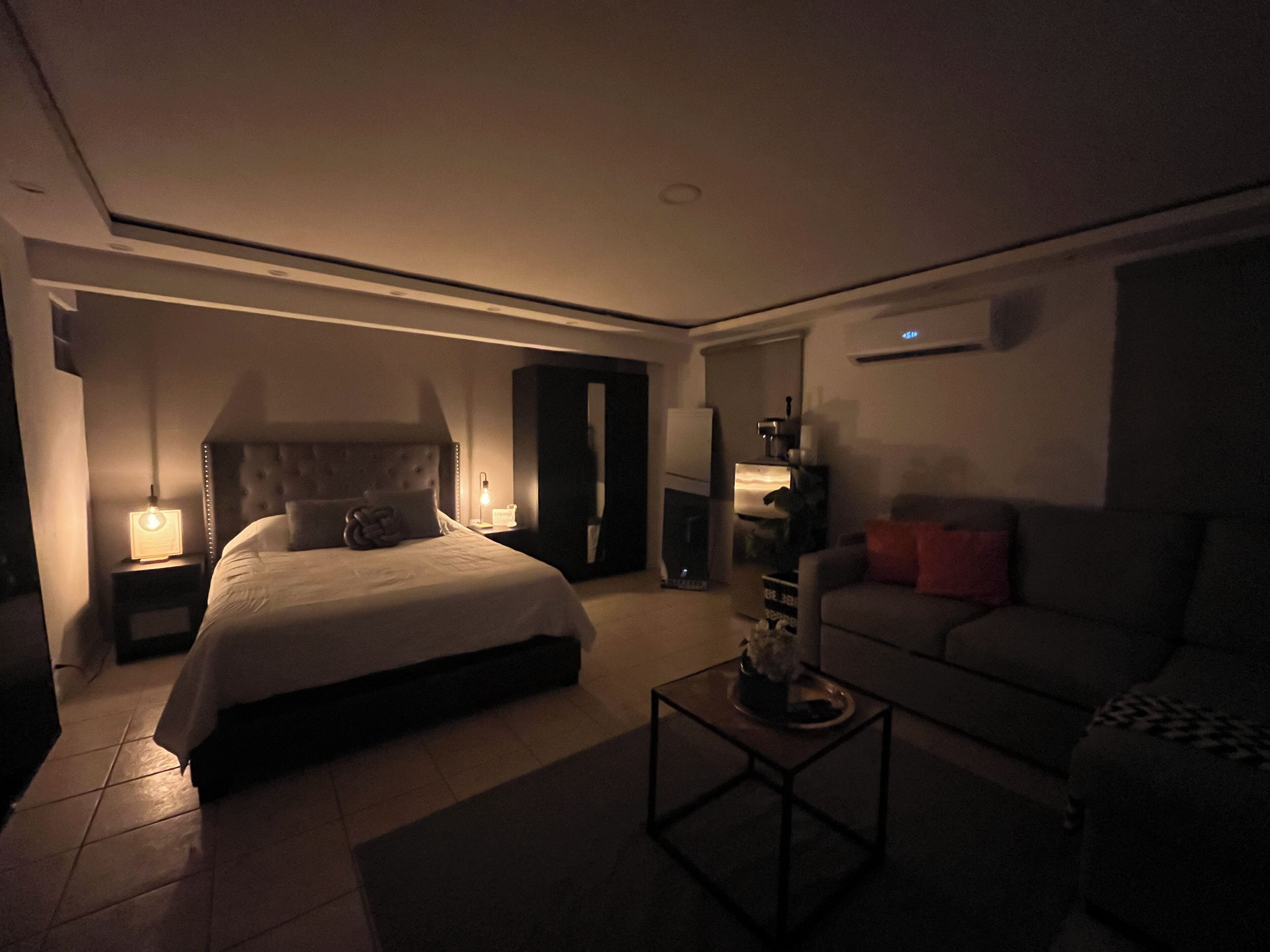Ein schwach beleuchtetes Schlafzimmer mit einem großen Bett, einem Nachttisch mit Lampe, einem Sofa und einem Couchtisch.