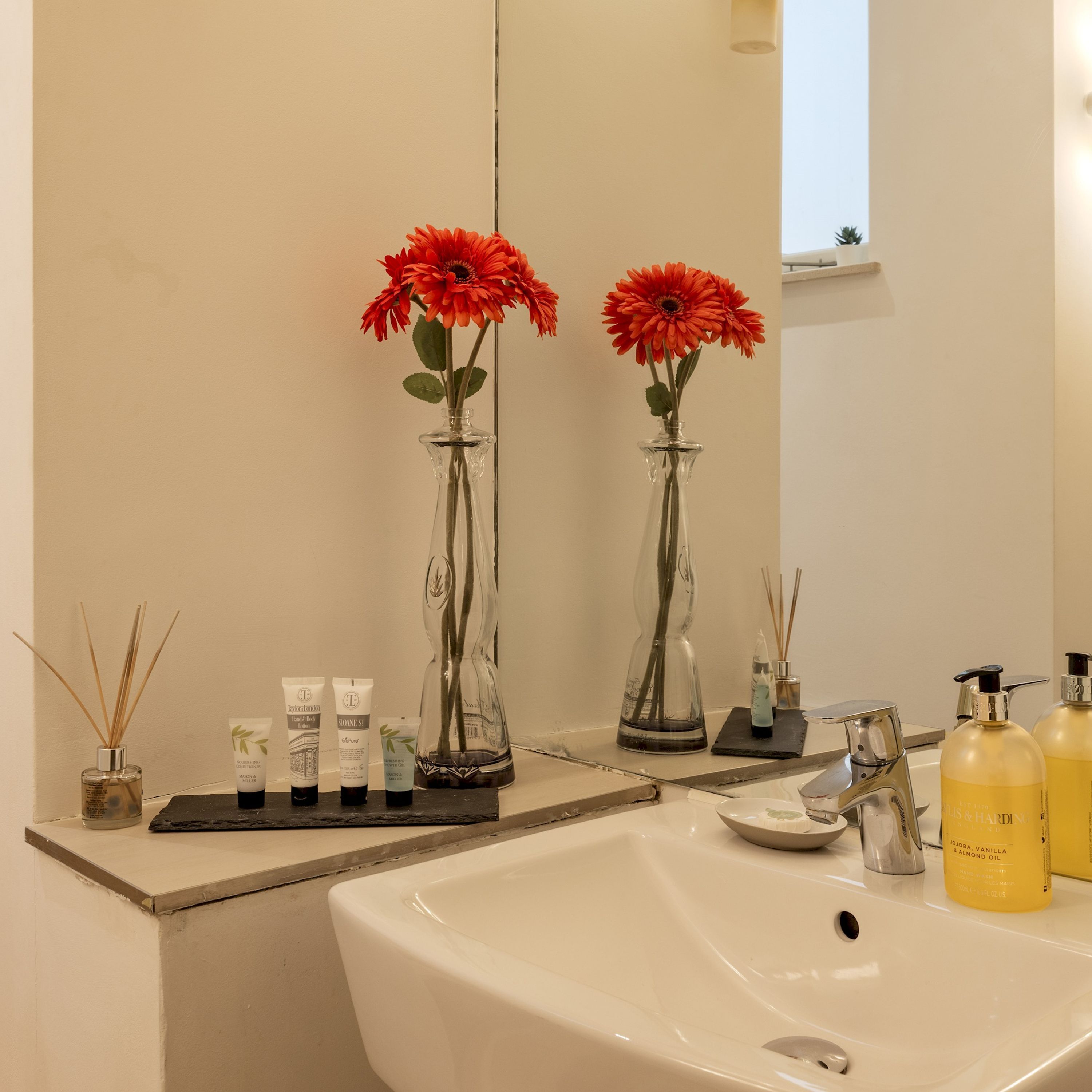 A bathroom with a large sink, two glass vases with red flowers, and toiletries on the counter.