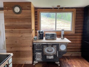 A rustic cabin interior with a wooden wall, a clock, a window with a view of a forest, a stove, and a toaster on a wooden counter.