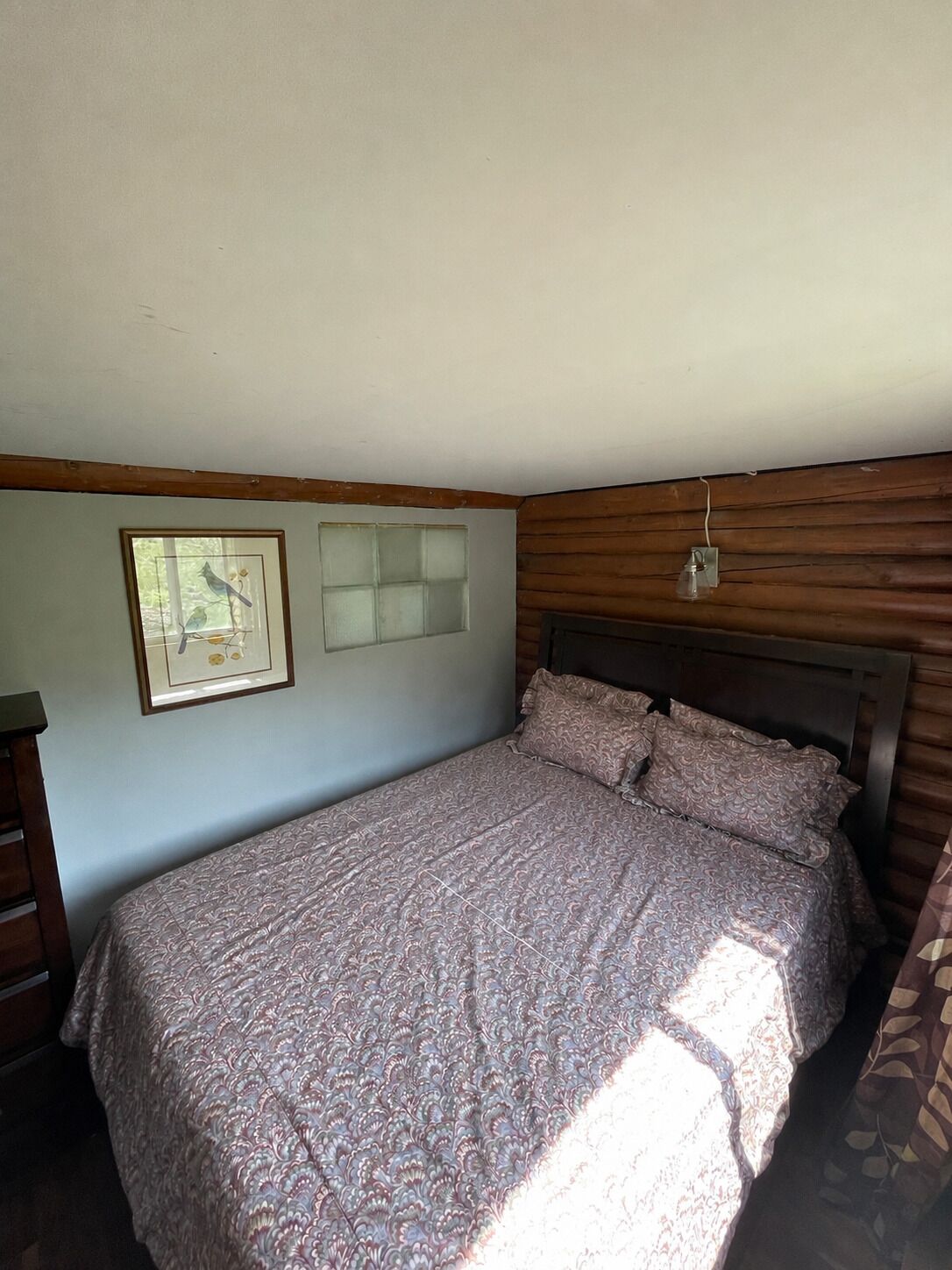 Un dormitorio con cama, un cuadro enmarcado, una ventana y una lámpara fijada a la pared.