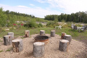 An outdoor seating area with a fire pit surrounded by wooden stumps, a table, and chairs. - At The River — Secluded Cabin with Private River Access (Woodlands County)