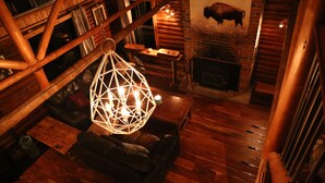 A cozy log cabin interior with a wooden ceiling, a fireplace, a hanging light fixture, and a couch.
