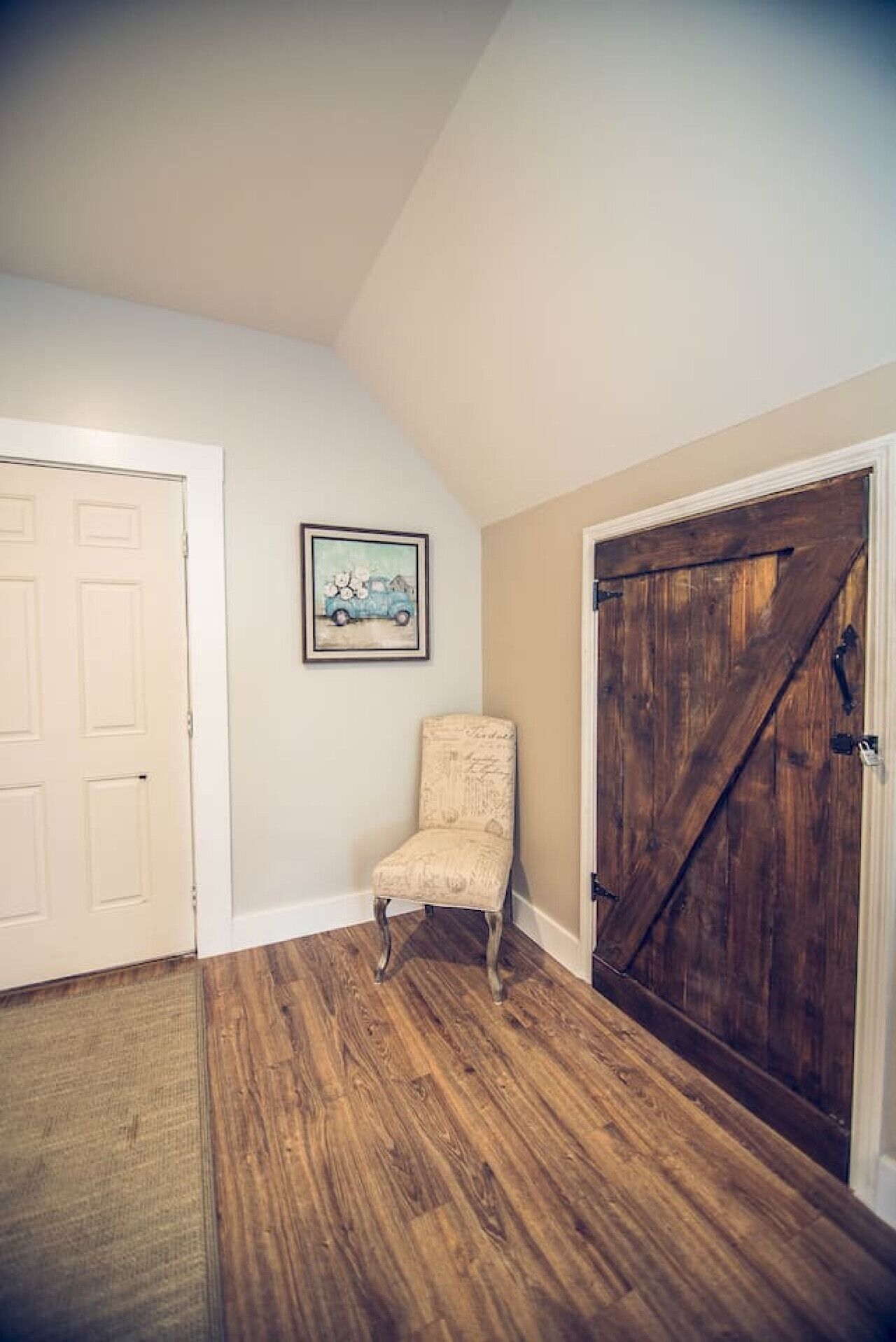 Una habitación con piso de madera, una puerta blanca, un cuadro en la pared y una silla.