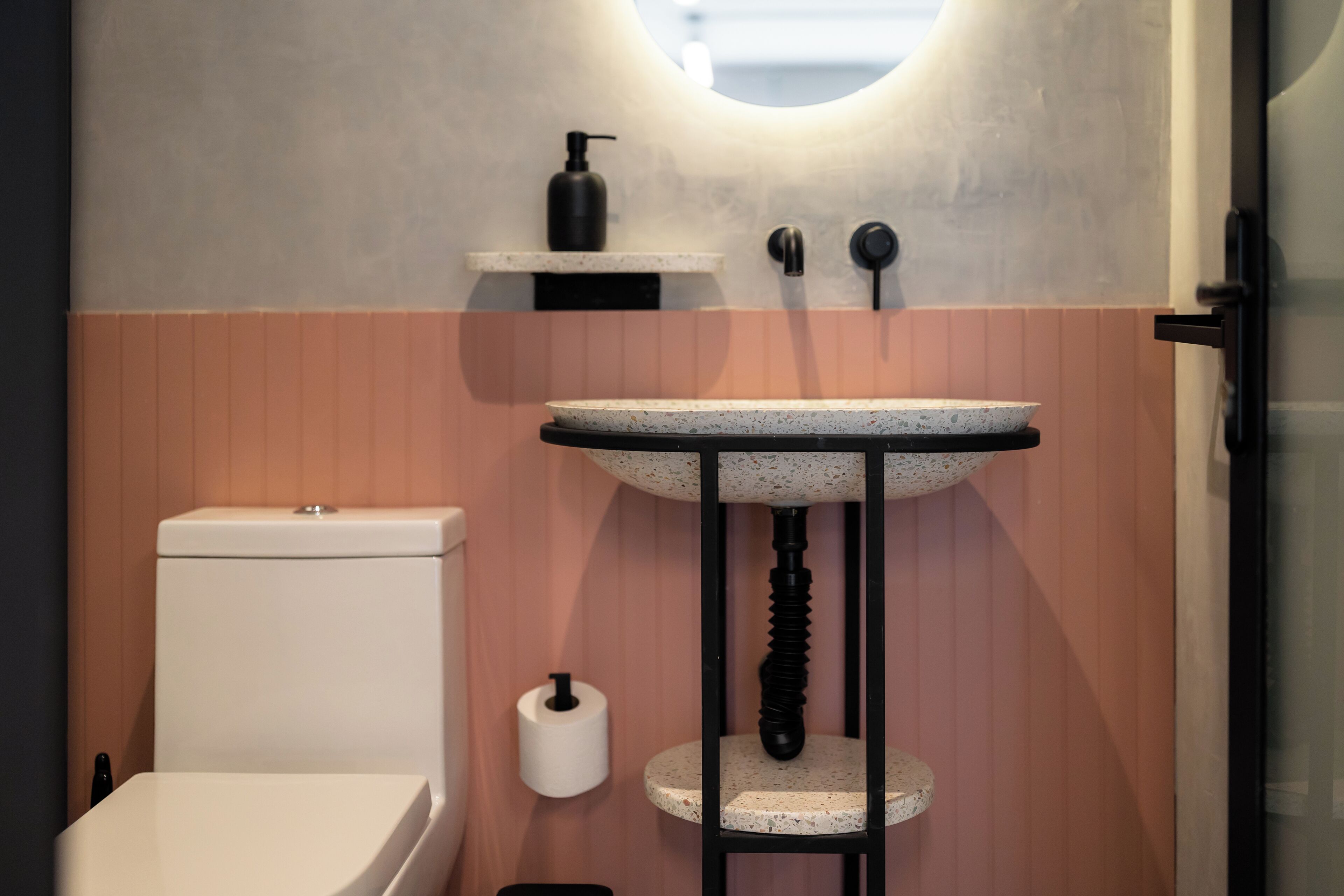 Un baño con un inodoro blanco, un lavamanos independiente con una pileta salpicada, un dispensador de jabón y un sujetarropas en la pared.