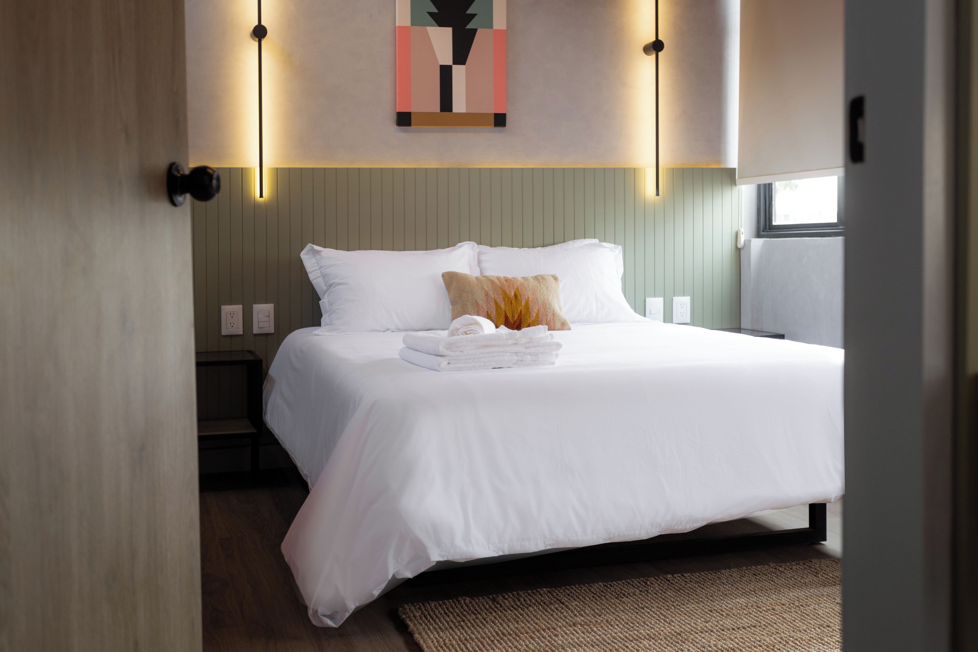 Una cama bien hecha con sábanas blancas y una toalla doblada, una lámpara de pared y una ventana con luz natural.