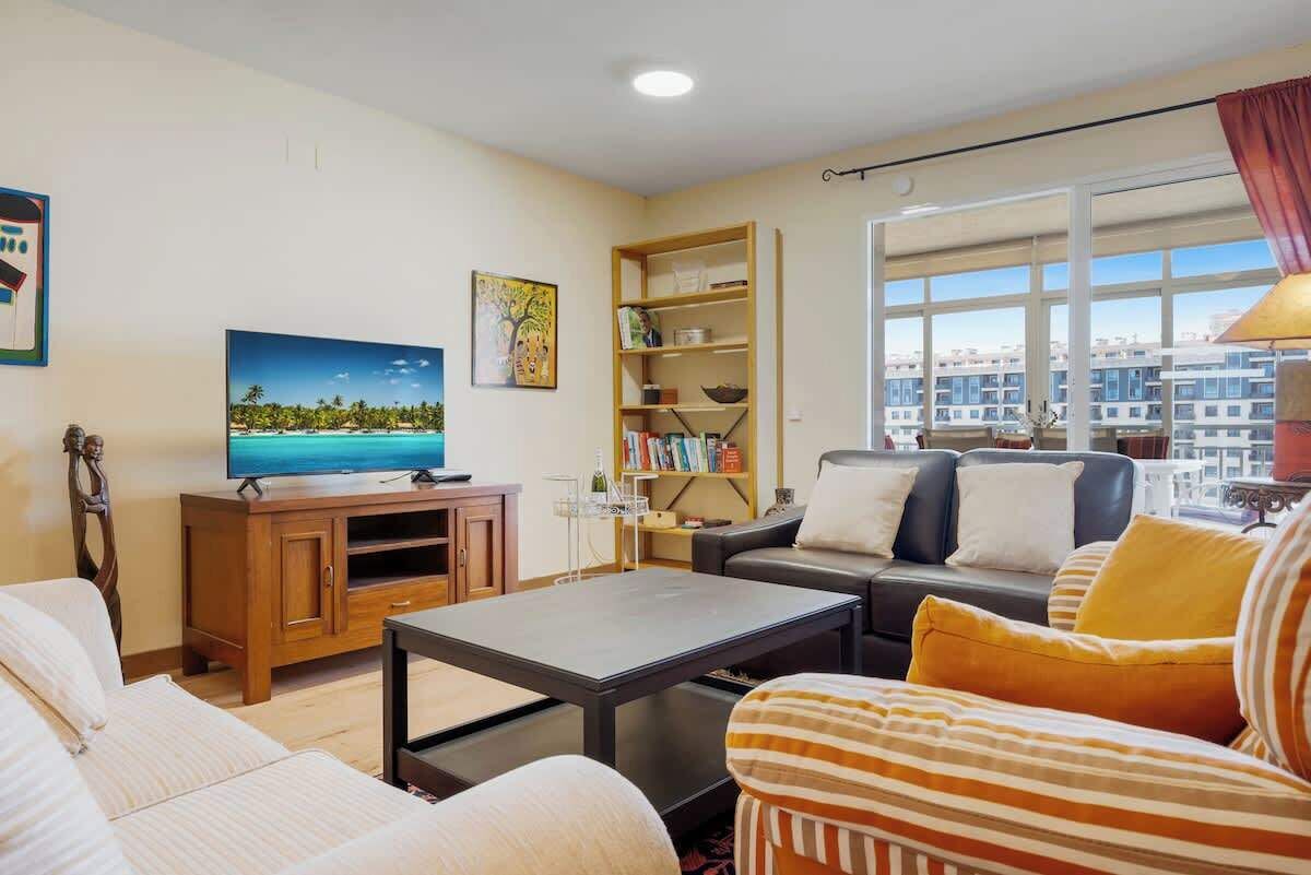 Een woonkamer met een televisie, bank, fauteuil, salontafel, boekenkast en grote ramen met uitzicht op de stad.