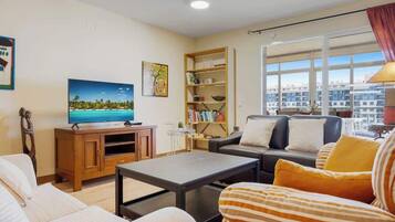 Een woonkamer met een televisie, bank, fauteuil, salontafel, boekenkast en grote ramen met uitzicht op de stad.