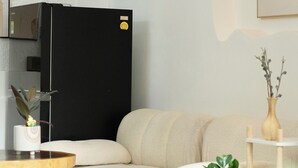 A modern living room with a beige sofa, a black refrigerator, a small wooden table, and a potted plant. - TRIP HOME stay & studio (Ho Chi Minh City)